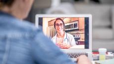 A female with a doctor's coat and glasses conducts a telehealth video call. There is a shoulder of a patient in the foreground.
